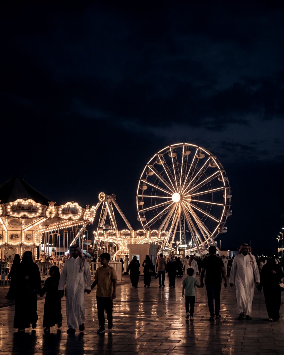A lit ferris wheel and amusement rides on the Jeddah corniche at night with families on the promenade