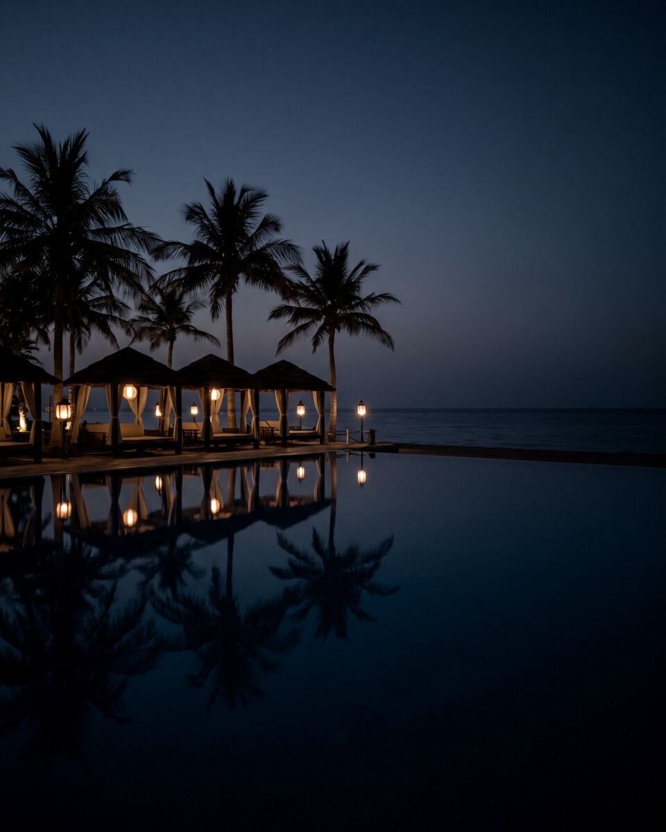 A quiet beach resort pool on the Obhur coast at blue hour, lit cabanas and palms reflected in the water
