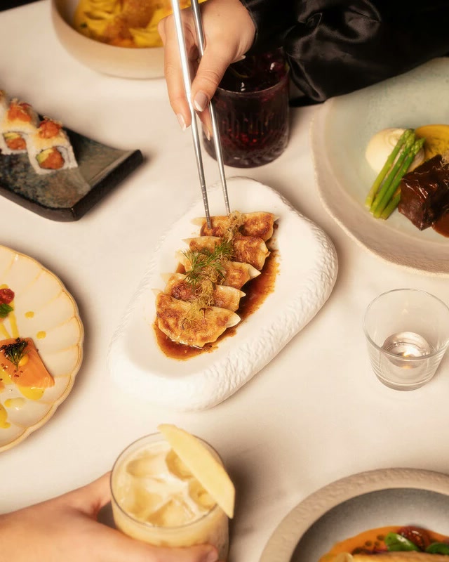 A dinner table from above: dumplings lifted with chopsticks, salmon crudo, sushi and slow cooked short rib