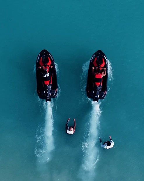 Two jet skis side by side over turquoise water, seen from above, with two swimmers waving them on